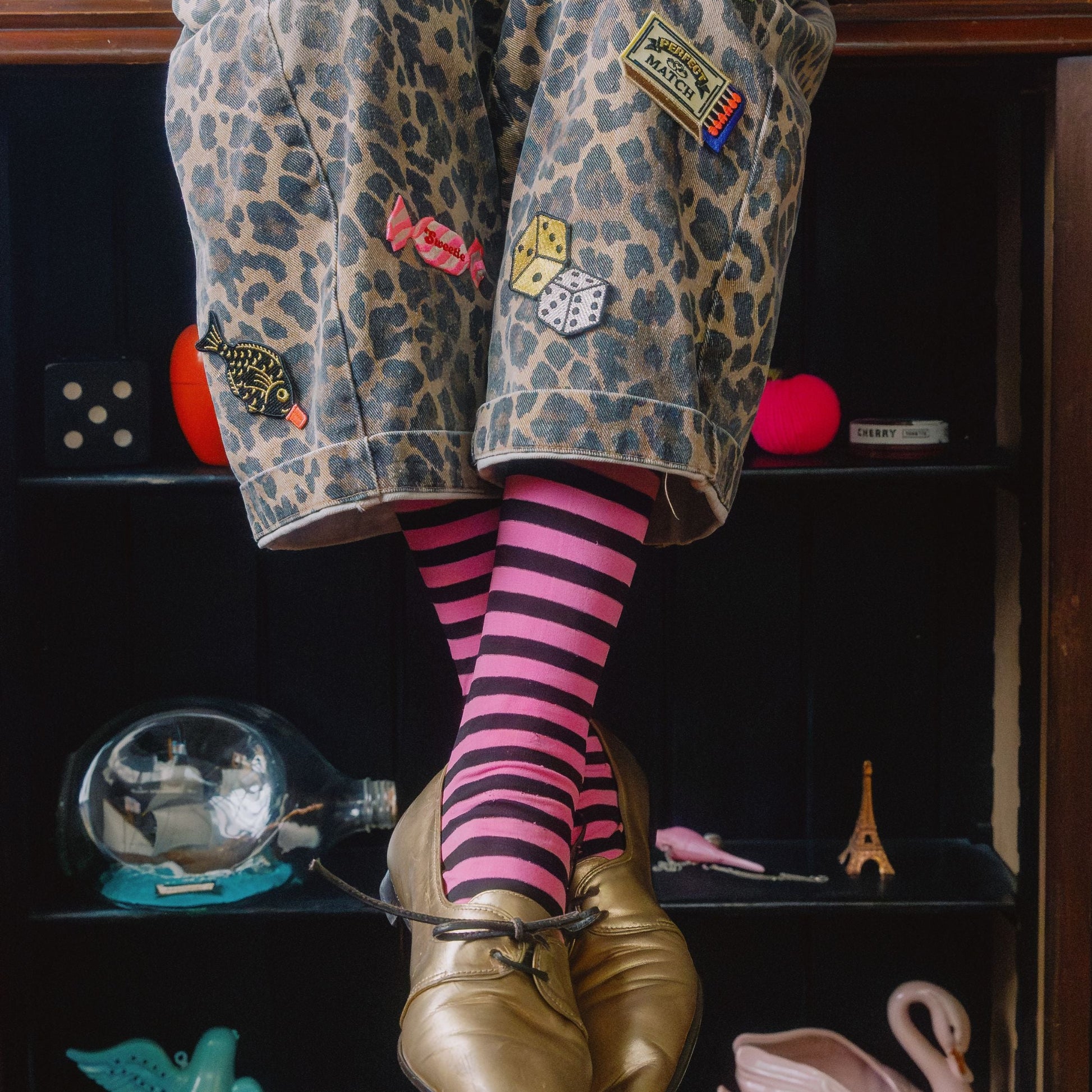 Person wearing leopard print pants with patches and pink striped socks, sitting on a shelf with decorative items.