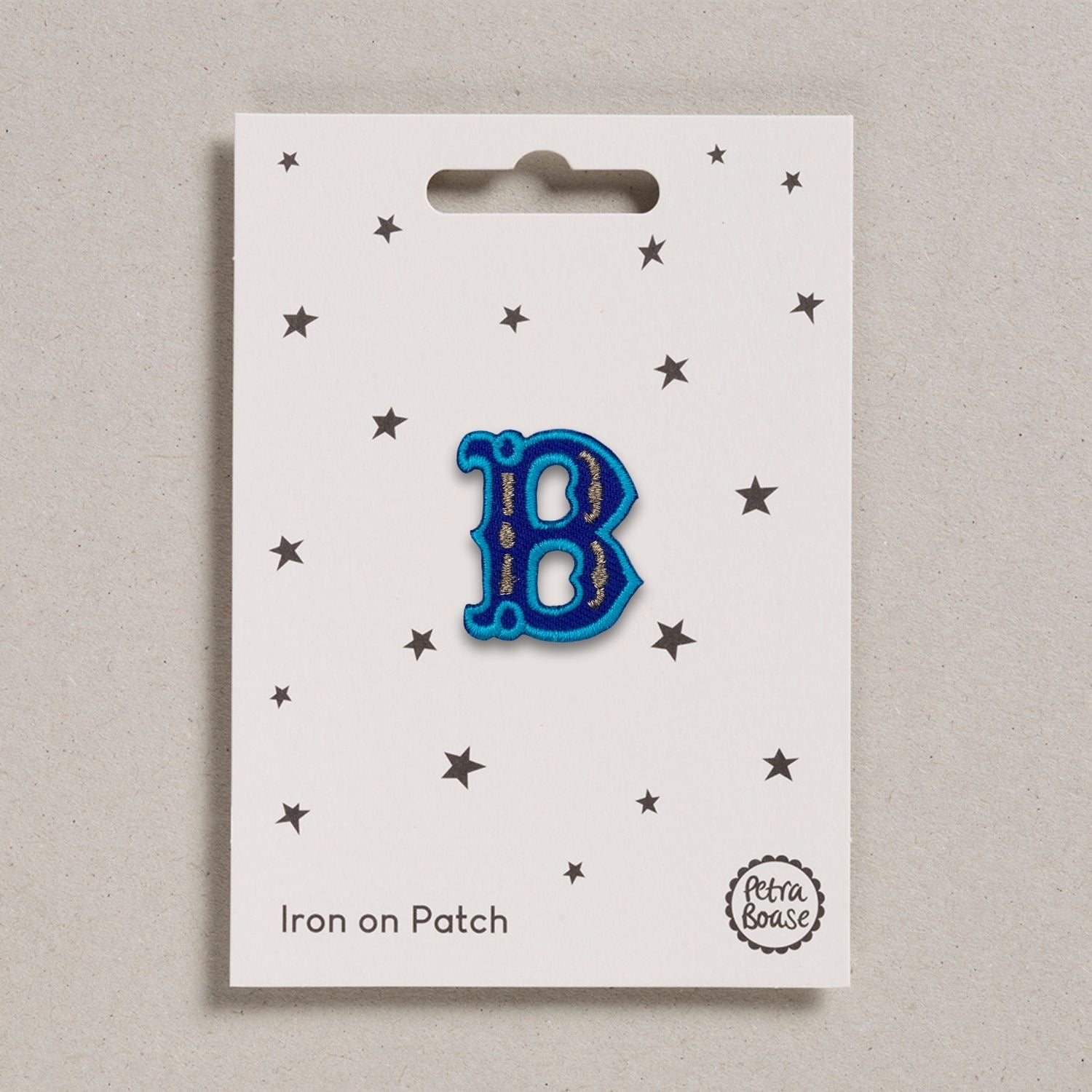 A blue embroidered letter 'B' patch with turquoise embroidered edging and brown metallic stitch detail, presented on a card with stars and 'Iron on Patch' text.