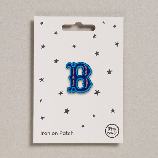 A blue embroidered letter 'B' patch with turquoise embroidered edging and brown metallic stitch detail, presented on a card with stars and 'Iron on Patch' text.