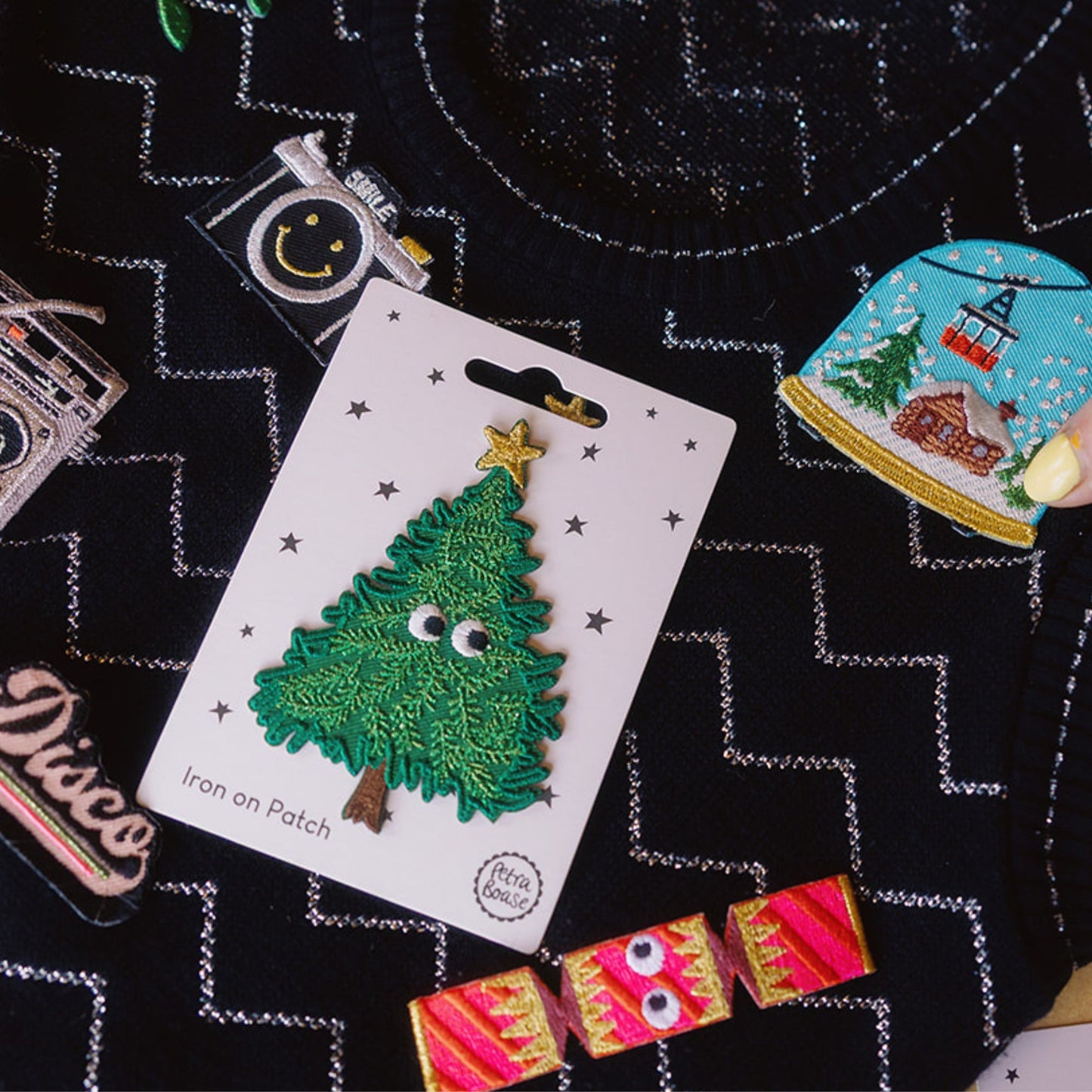 Christmas tree iron-on patch on a white card with other decorative patches on a black sparkly vintage jumper.