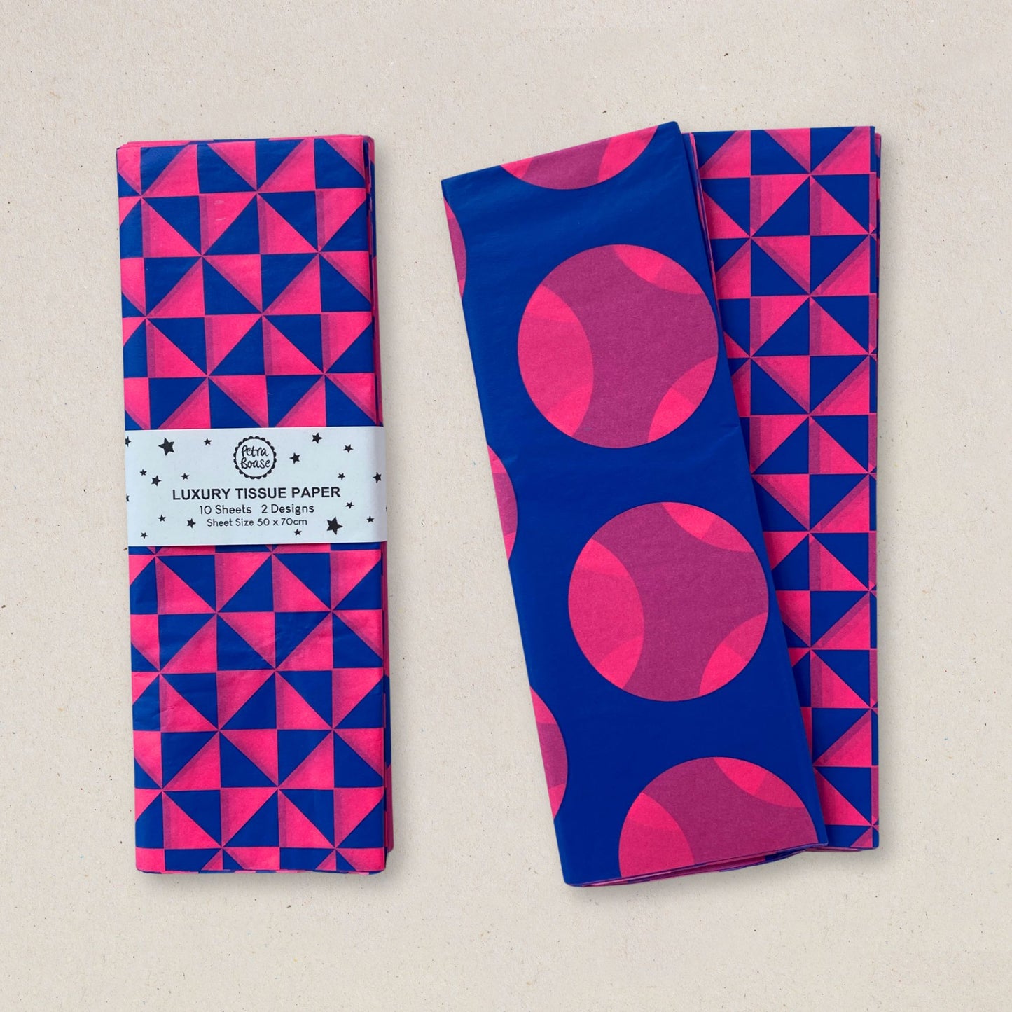 Two folded pieces of patterned tissue paper on a light beige background. One has a blue and pink geometric triangle pattern with a paper band label reading 'Luxury Tissue Paper'; the other has a blue and pink geometric triangle pattern with large pink circles.