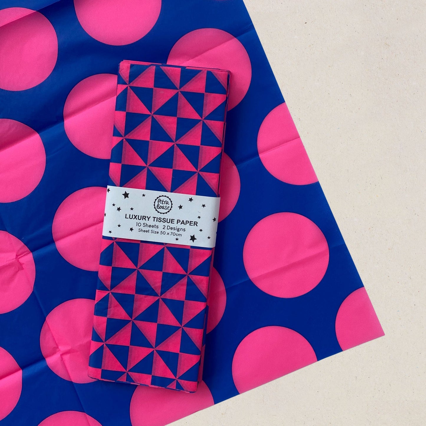 A folded roll of patterned tissue paper with a white paper band label reading 'LUXURY TISSUE PAPER' placed on top of a larger matching sheet with pink polka dots on a blue background.