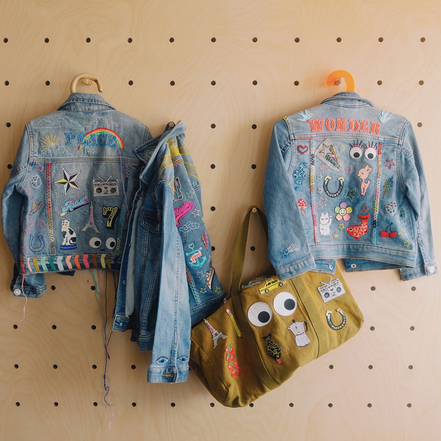 Three denim jackets and a matching olive tote bag covered in colorful embroidered patches and appliqués, hanging on a pegboard wall.