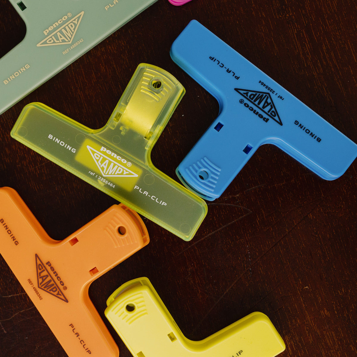 Several colorful plastic binder clips from Penco PLA Clip collection are arranged on a dark wooden surface, next to a yellow zipper pouch.