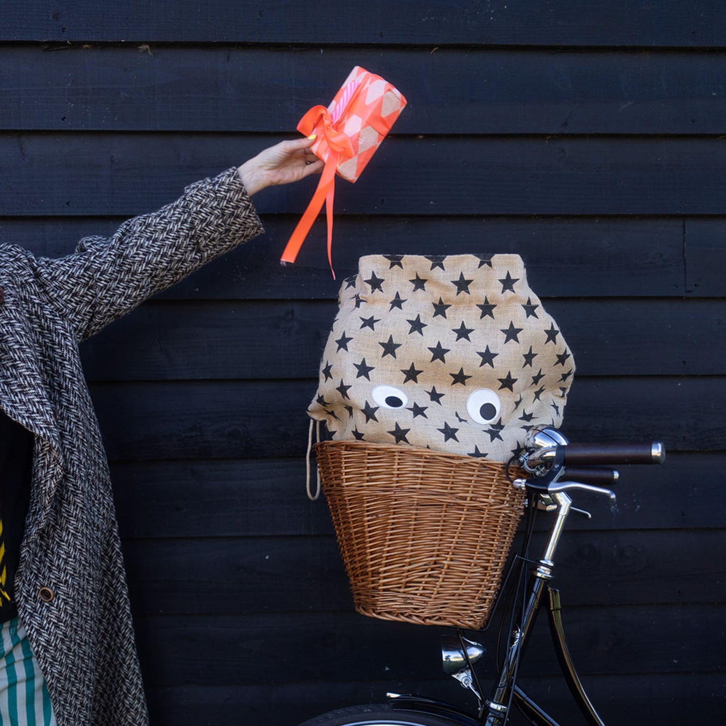 Person holding a gift above a bicycle basket with a star pattern jute Christmas sack against a dark wooden wall.