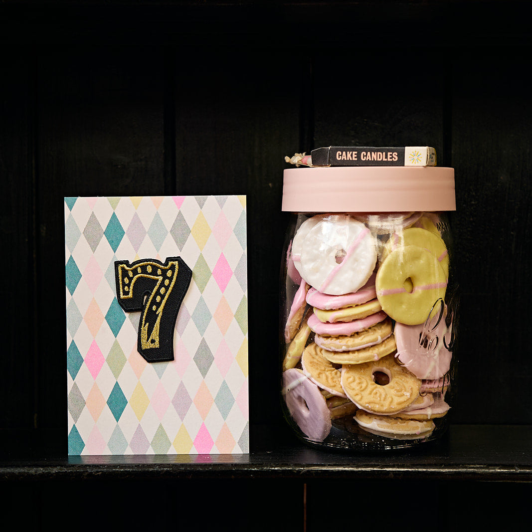 Jar of colorful cake candles with a birthday card featuring the number 7 on a black shelf.
