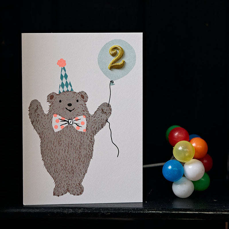 Card with a bear holding a balloon with the number 2 on a black surface.