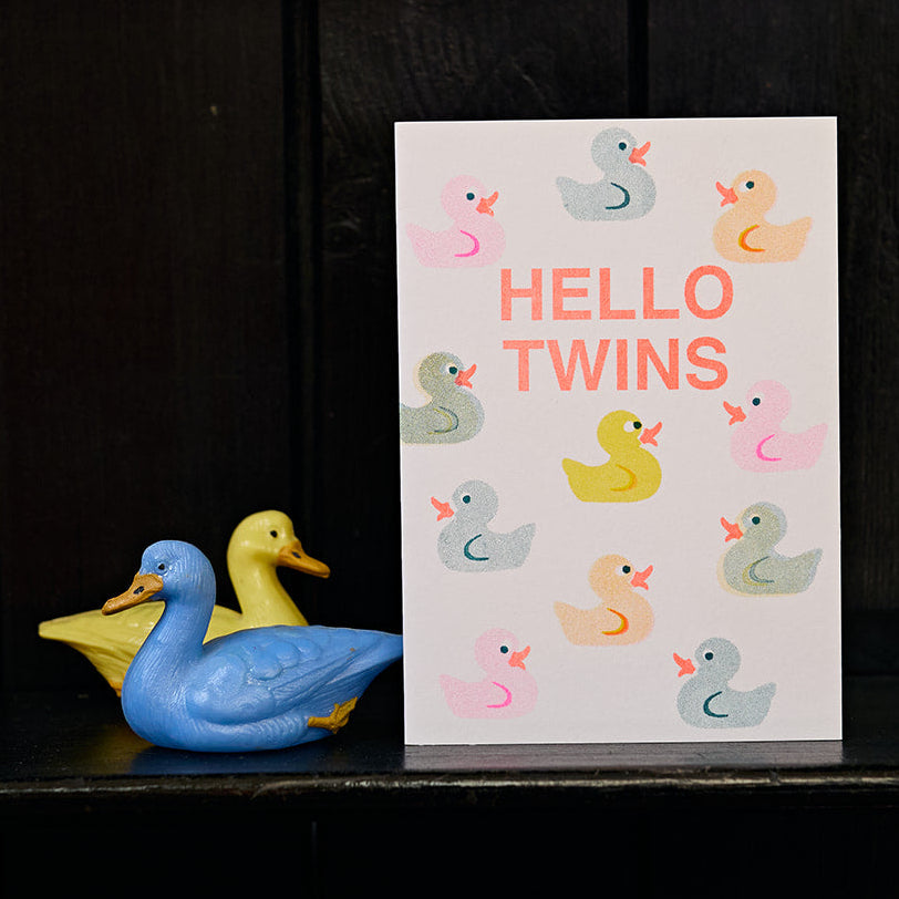 Card with 'Hello Twins' text and duck illustrations on a dark surface with toy ducks.