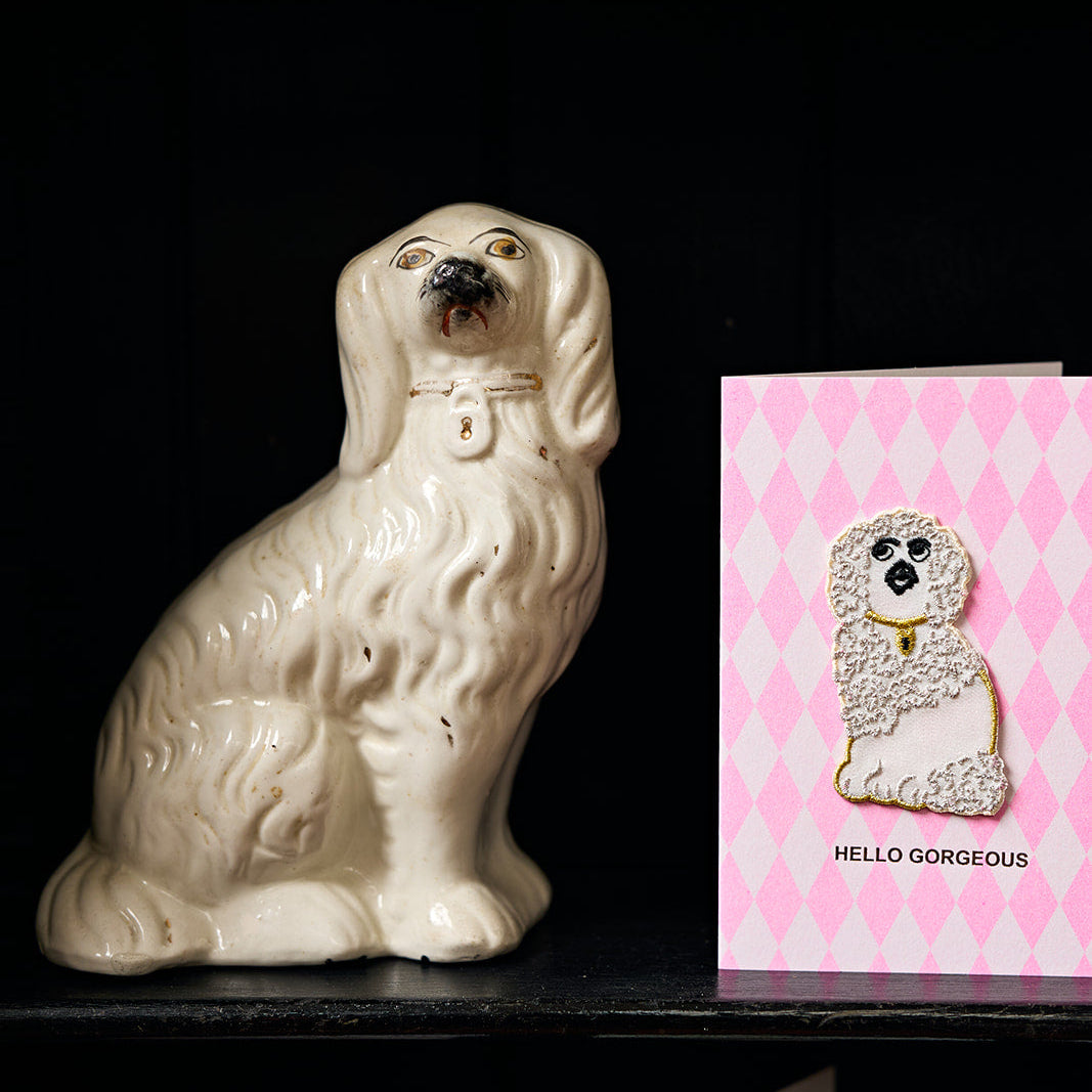 White ceramic dog figurine next to a pink card with a dog illustration on a black shelf.