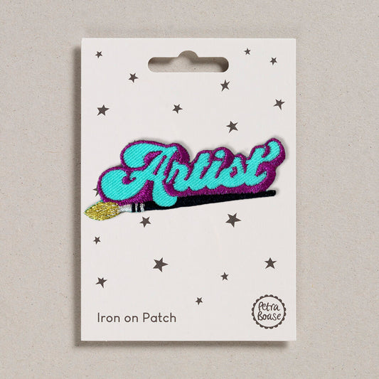 A packaged 'Artist' iron on patch with blue, purple, black, and gold embroidered detailing, displayed on a card with stars.