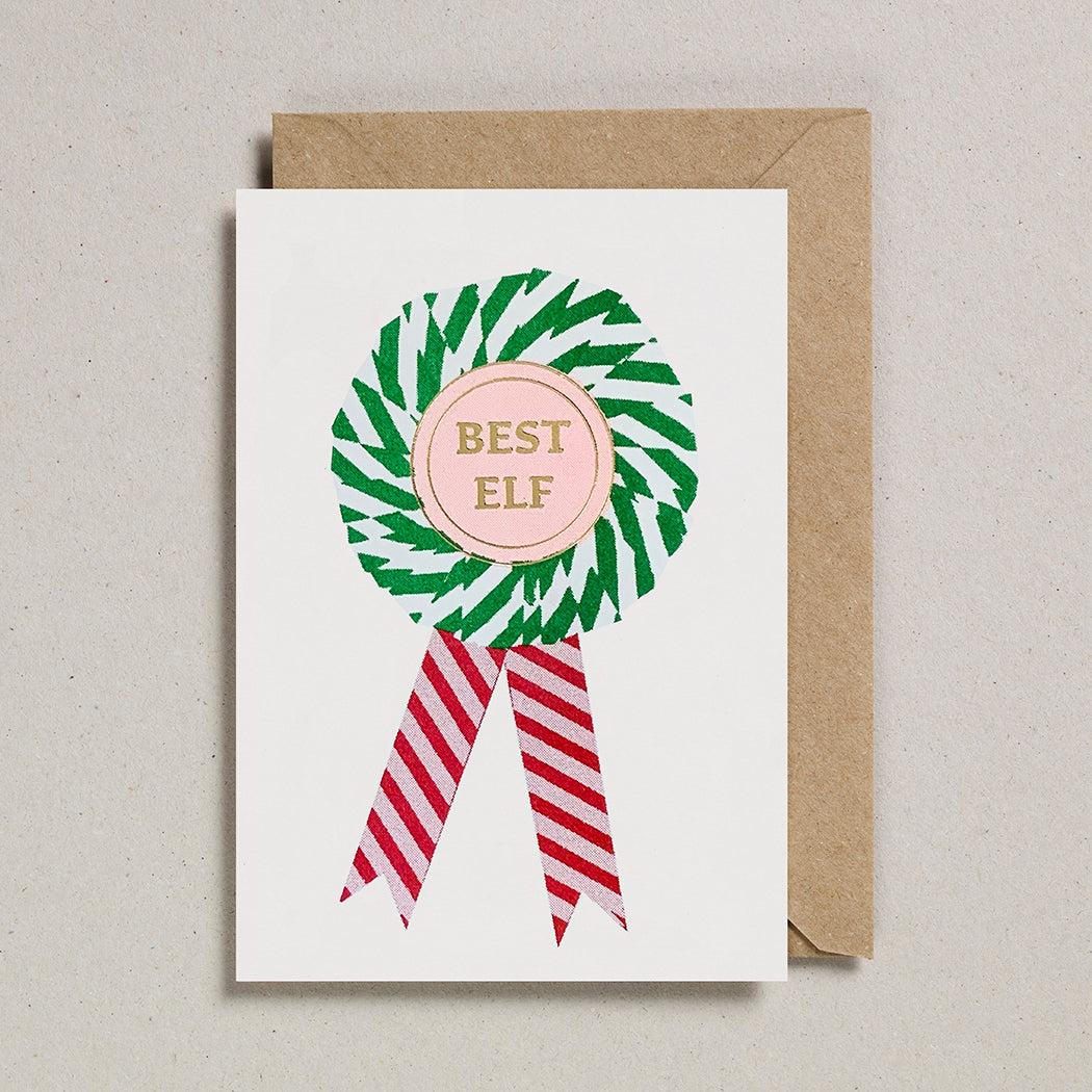 A greeting card with a risograph print of a rosette award with 'BEST ELF' written in the center, using soy-based inks on FSC certified card, embellished with a foiled emblem, with a red and white striped ribbon, and a brown envelope.