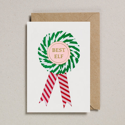 A greeting card with a risograph print of a rosette award with 'BEST ELF' written in the center, using soy-based inks on FSC certified card, embellished with a foiled emblem, with a red and white striped ribbon, and a brown envelope.
