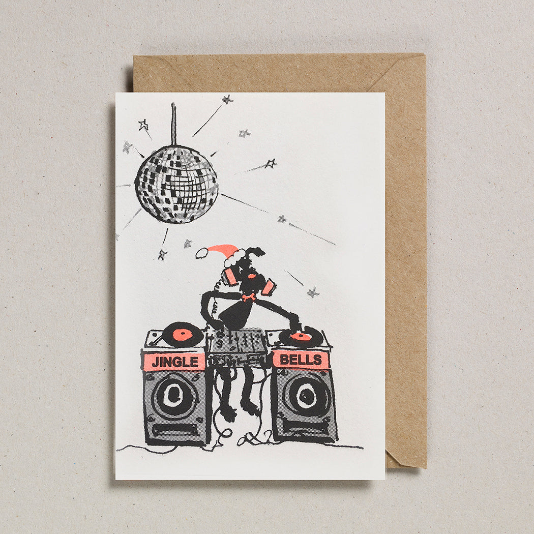 A greeting card with a black and white illustration of a character DJing with a disco ball and equipment, titled 'JINGLE BELLS'. Accompanied by a brown envelope.